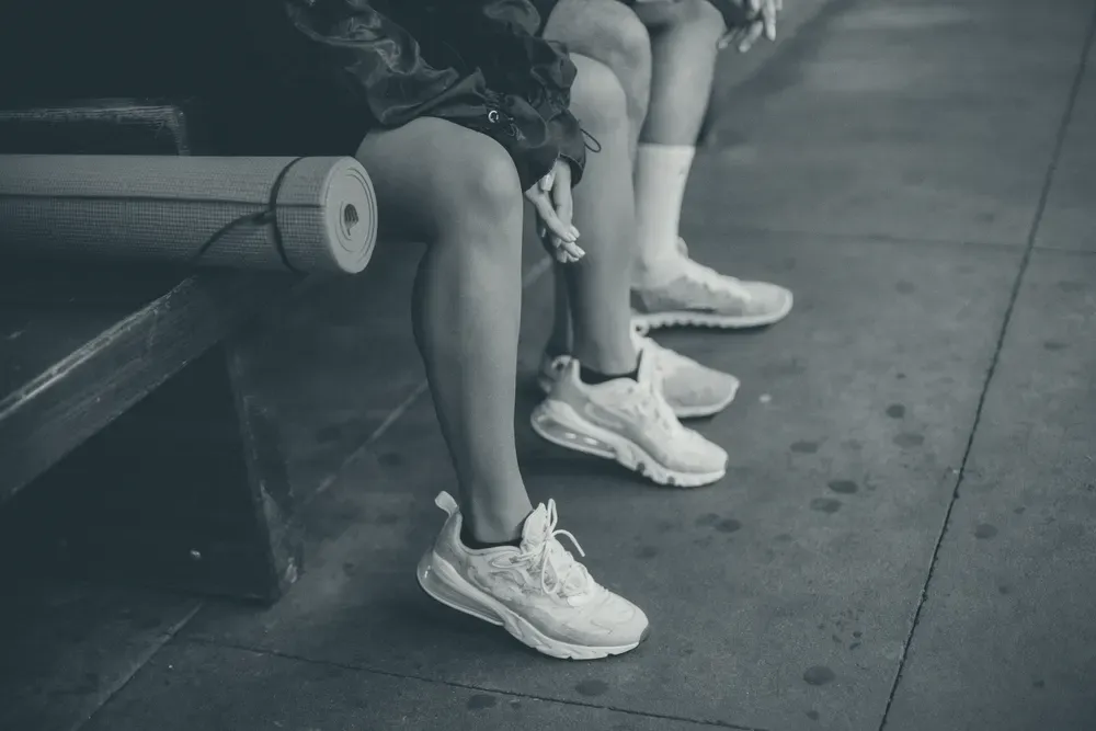 Pernas de duas pessoas sentadas em um banco de madeira usando tênis de corrida, com um tapete de exercícios enrolado ao lado.