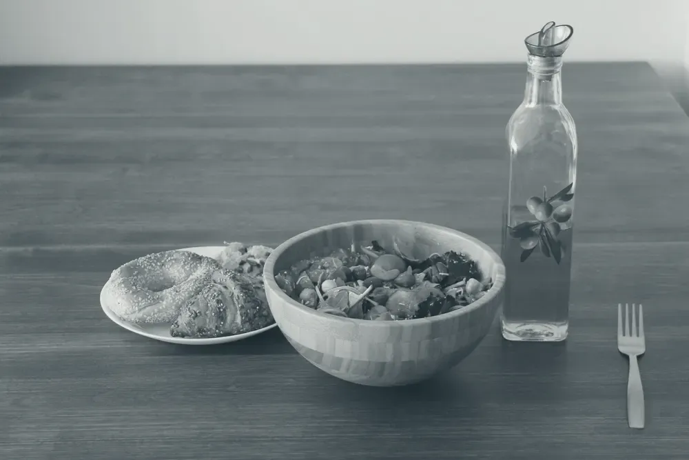 Tigela de madeira com salada de folhas verdes ao lado de uma garrafa de azeite de oliva sobre a mesa.