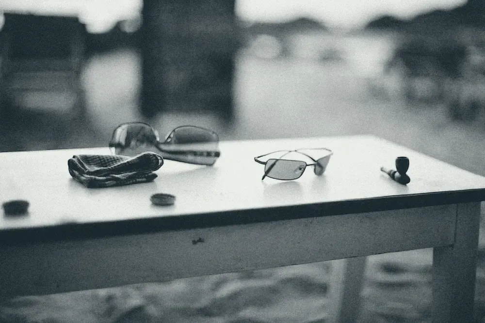 Óculos escuros e óculos de grau apoiados sobre uma mesa, em preto e branco.