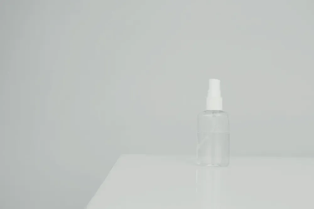 Frasco spray transparente sobre uma mesa branca, com parede em tom bege ao fundo