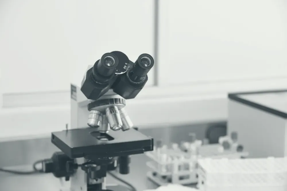 Microscópio de laboratório em primeiro plano, com tubos de amostra de sangue desfocados ao fundo.