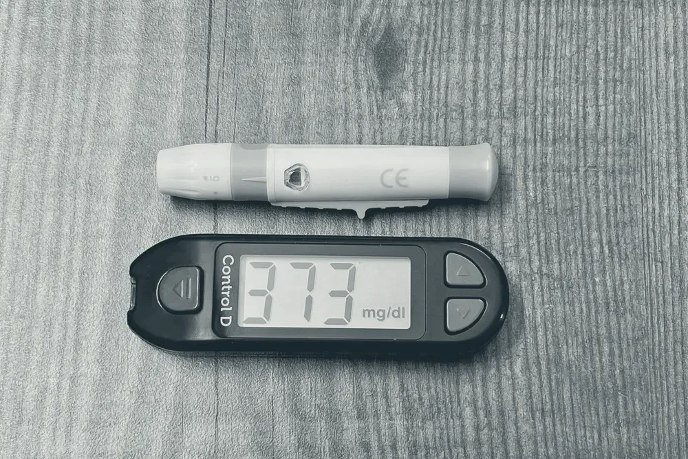 Glicosímetro digital exibindo uma leitura de glicemia ao lado de uma caneta de lanceta sobre superfície de madeira.