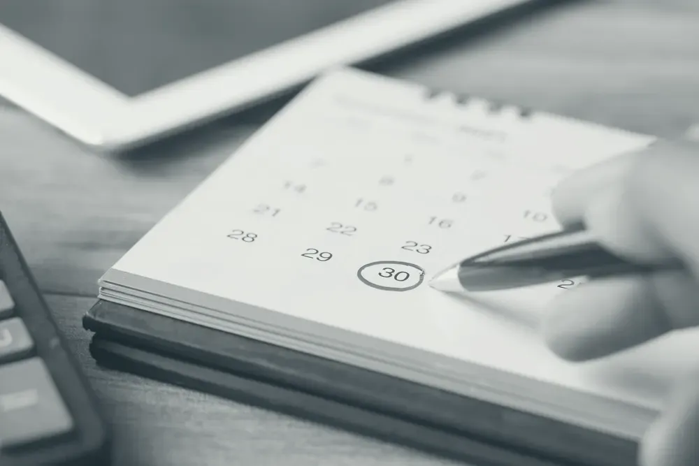 Mão segurando uma caneta sobre um calendário de mesa, com um dia circulado em vermelho